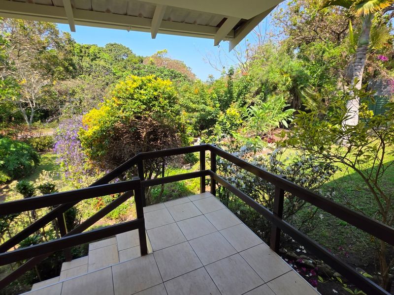 A covered outdoor patio featuring tiled flooring and a timber balustrade, overlooking a lush, established garden.