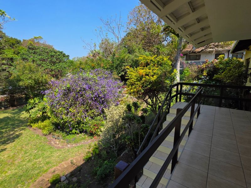A covered patio featuring tiled flooring and a timber balustrade, overlooking a lush, well-established garden with mature trees and shrubs.