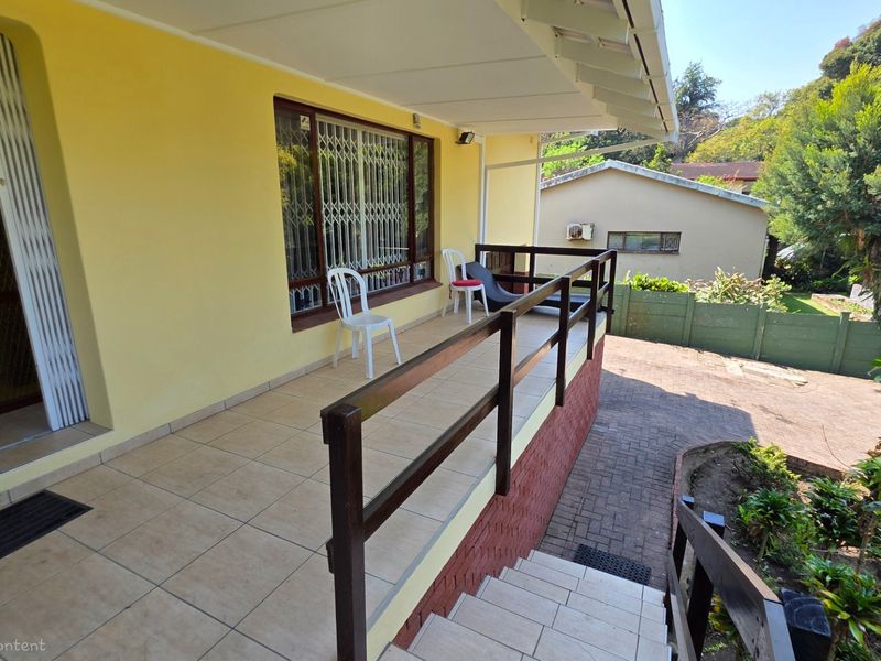 A covered patio area featuring tiled flooring and a timber balustrade, providing an outdoor space overlooking the garden and driveway.