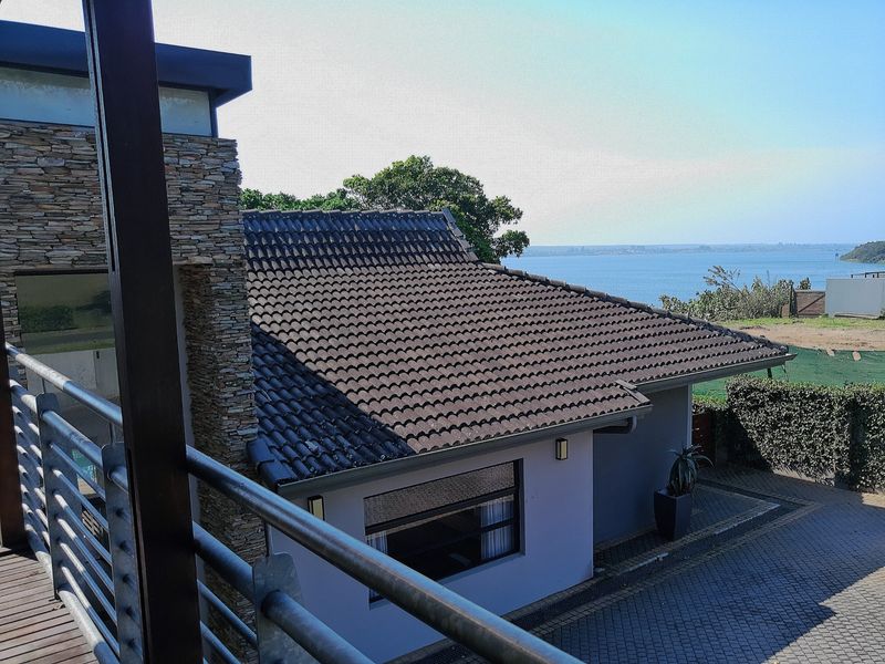 Elevated view of the rear exterior featuring a tiled roof, stone cladding, paved driveway, and a balcony overlooking the water.