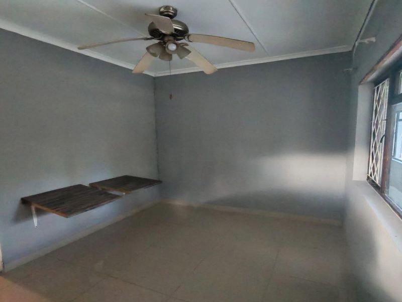 An empty bedroom featuring tiled flooring, a ceiling fan, and wall-mounted shelving, with a view into the adjacent kitchen area.