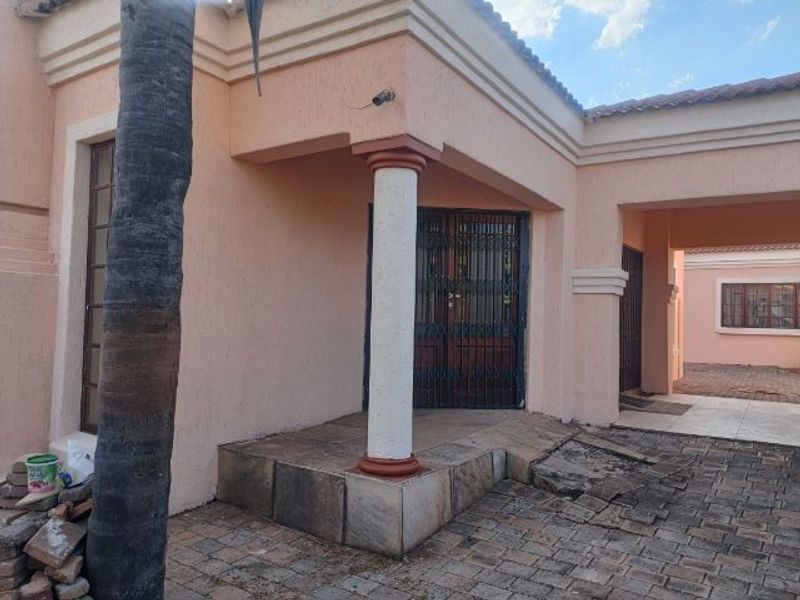 Front entrance area featuring a covered patio, security gate, and paved driveway, showing signs of wear and maintenance requirements on the paving and stone cladding.