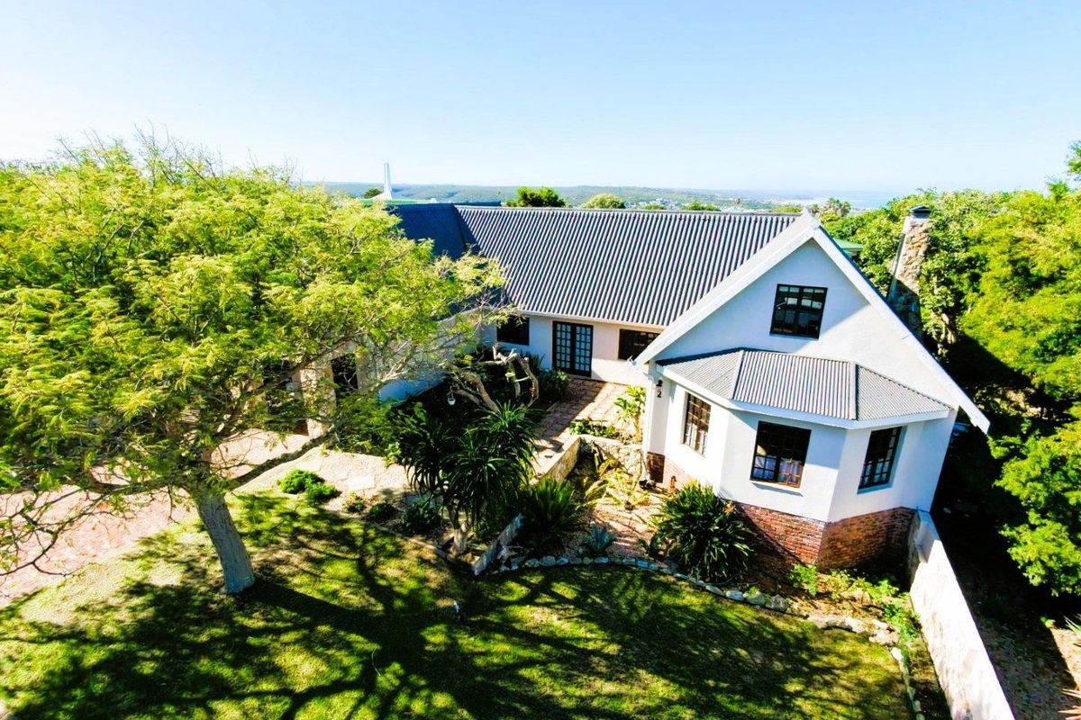 An elevated aerial view of a well-maintained residential property featuring a metal roof, stone chimney, and established garden landscaping.