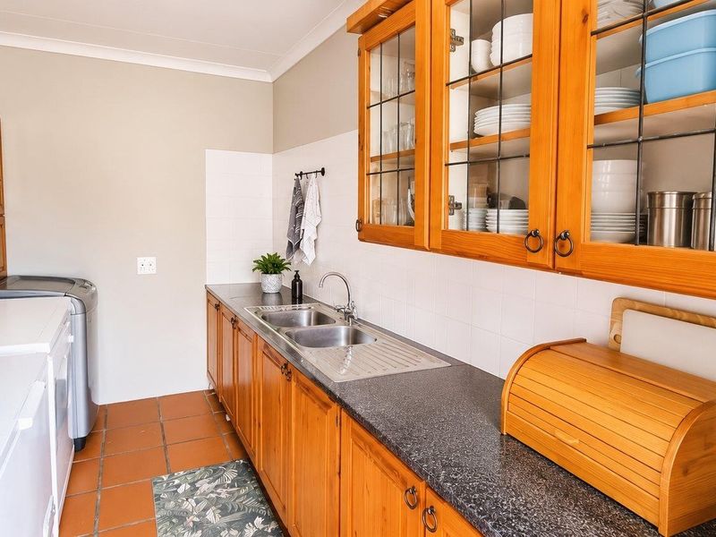 A functional scullery featuring timber cabinetry, double stainless steel sink, laminate countertops, and terracotta floor tiles, providing ample storage and prep space.