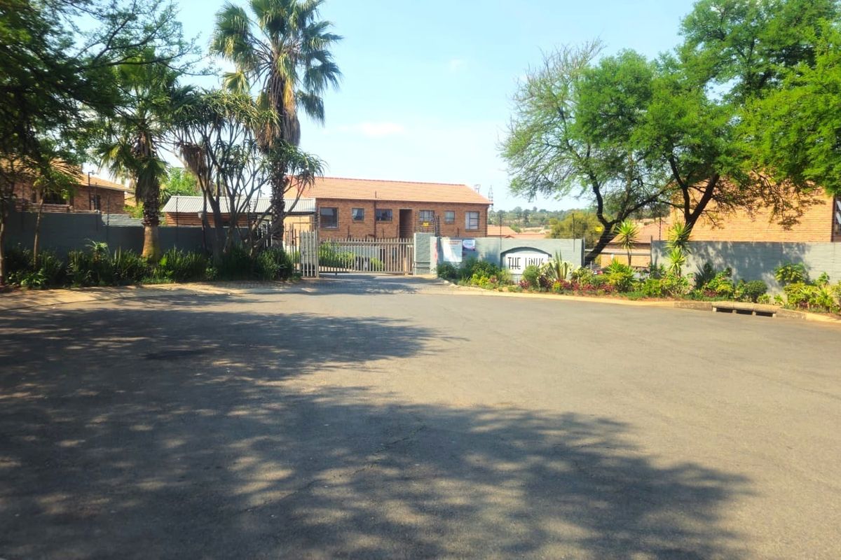 Street view of the apartment complex entrance featuring a paved driveway, perimeter wall, and security gate access.