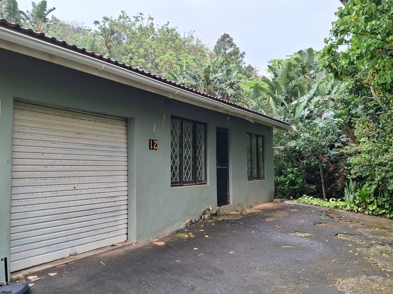 Exterior view of a townhouse unit featuring a roller shutter garage door, security-barred windows, and a weathered asphalt driveway surrounded by dense vegetation.