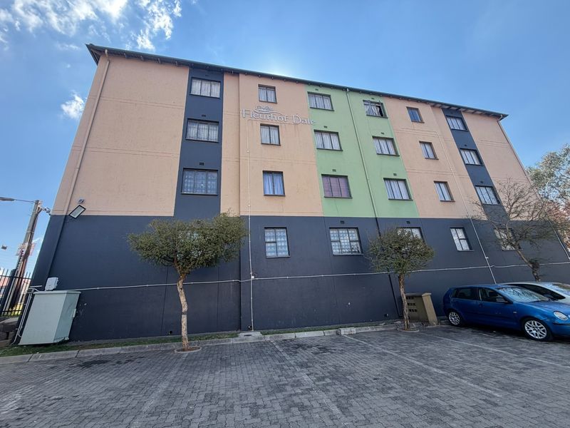 Exterior view of the Fleurhof Dale apartment complex featuring a multi-colored facade, security-barred windows, and an adjacent paved parking area.