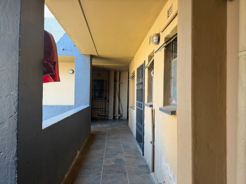 An exterior apartment walkway featuring tiled flooring, security gates, and wall-mounted lighting, showing signs of wear on the lower wall surfaces.