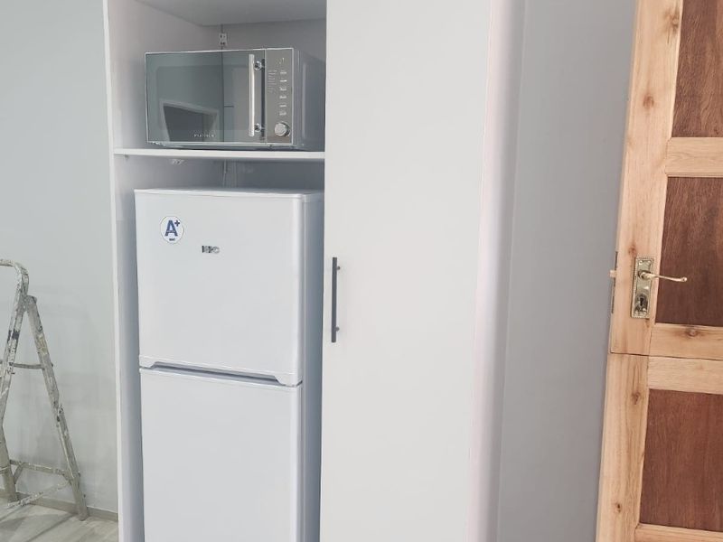 A compact kitchen area featuring white built-in cabinetry with dedicated space for a microwave and refrigerator, complemented by light-toned laminate flooring.