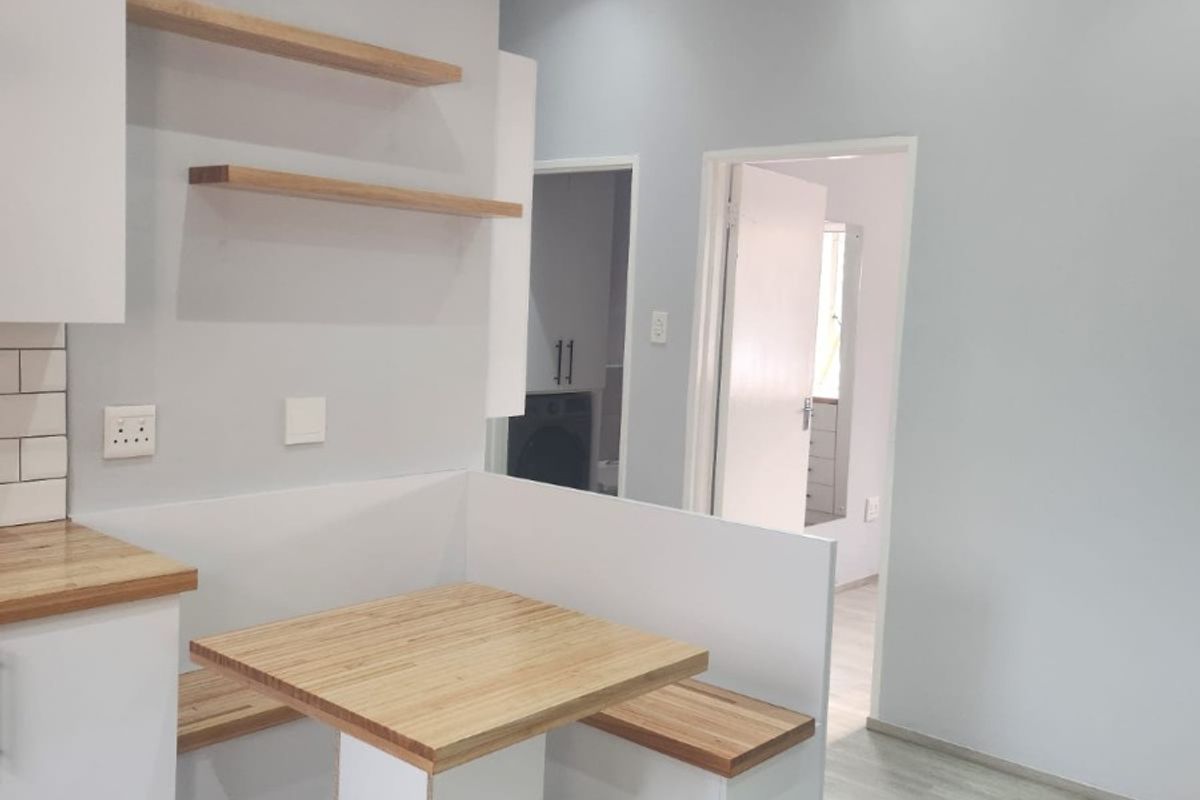 Modern kitchen area featuring a built-in breakfast nook with timber seating and table, floating shelves, and light-toned laminate flooring.