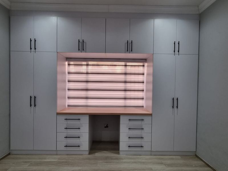 A modern bedroom featuring extensive white built-in cabinetry with an integrated desk area, light wood-look laminate flooring, and contemporary window blinds.