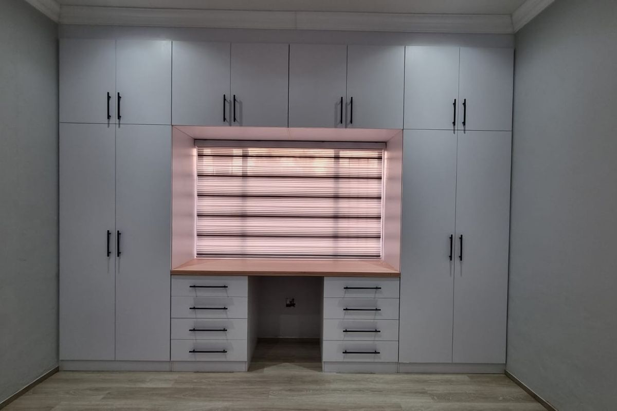 A modern bedroom featuring extensive white built-in cabinetry with an integrated desk area, light wood-look laminate flooring, and contemporary window blinds.