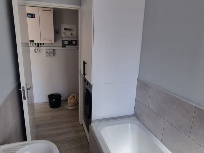 A clean bathroom featuring a pedestal basin, built-in bathtub, and tiled splashbacks, with an adjacent utility cupboard housing an inverter and electrical distribution board.