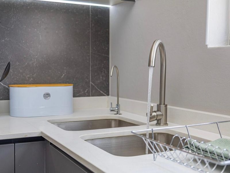 Modern kitchen area featuring a double stainless steel sink, gooseneck faucet, filtered water tap, stone countertops, and a dark stone splashback with integrated lighting.