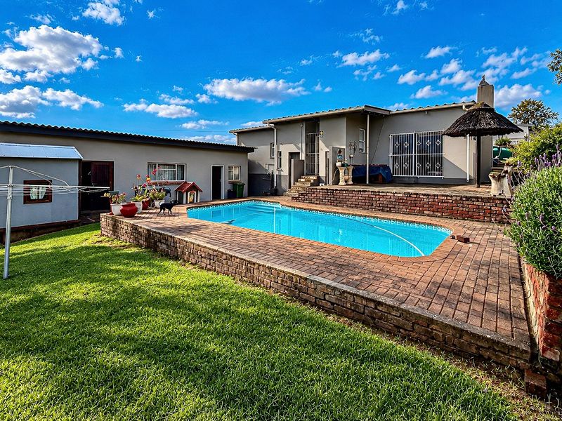 A backyard area featuring a swimming pool with brick paving, a raised brick retaining wall, and a lawn area adjacent to the main house structure.