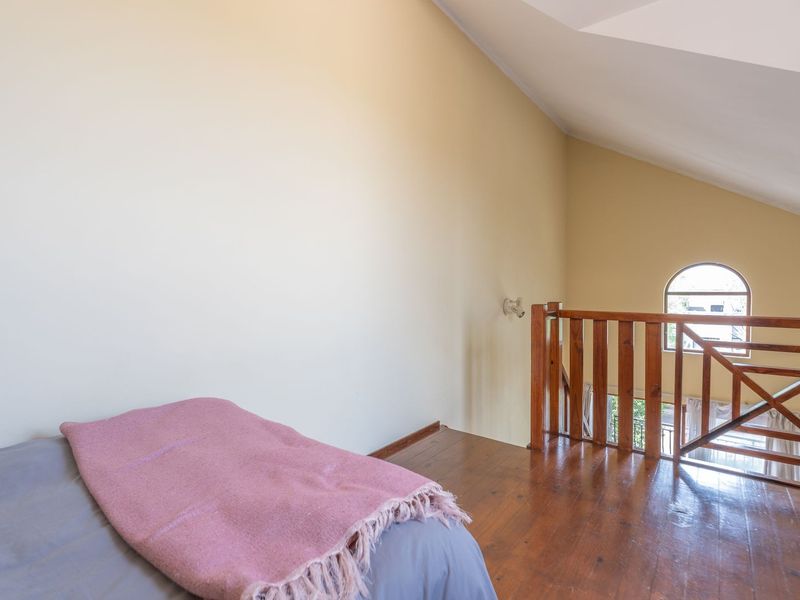 A loft-style bedroom featuring warm wooden flooring, a vaulted ceiling, and a wooden balustrade overlooking the lower level through an arched window.