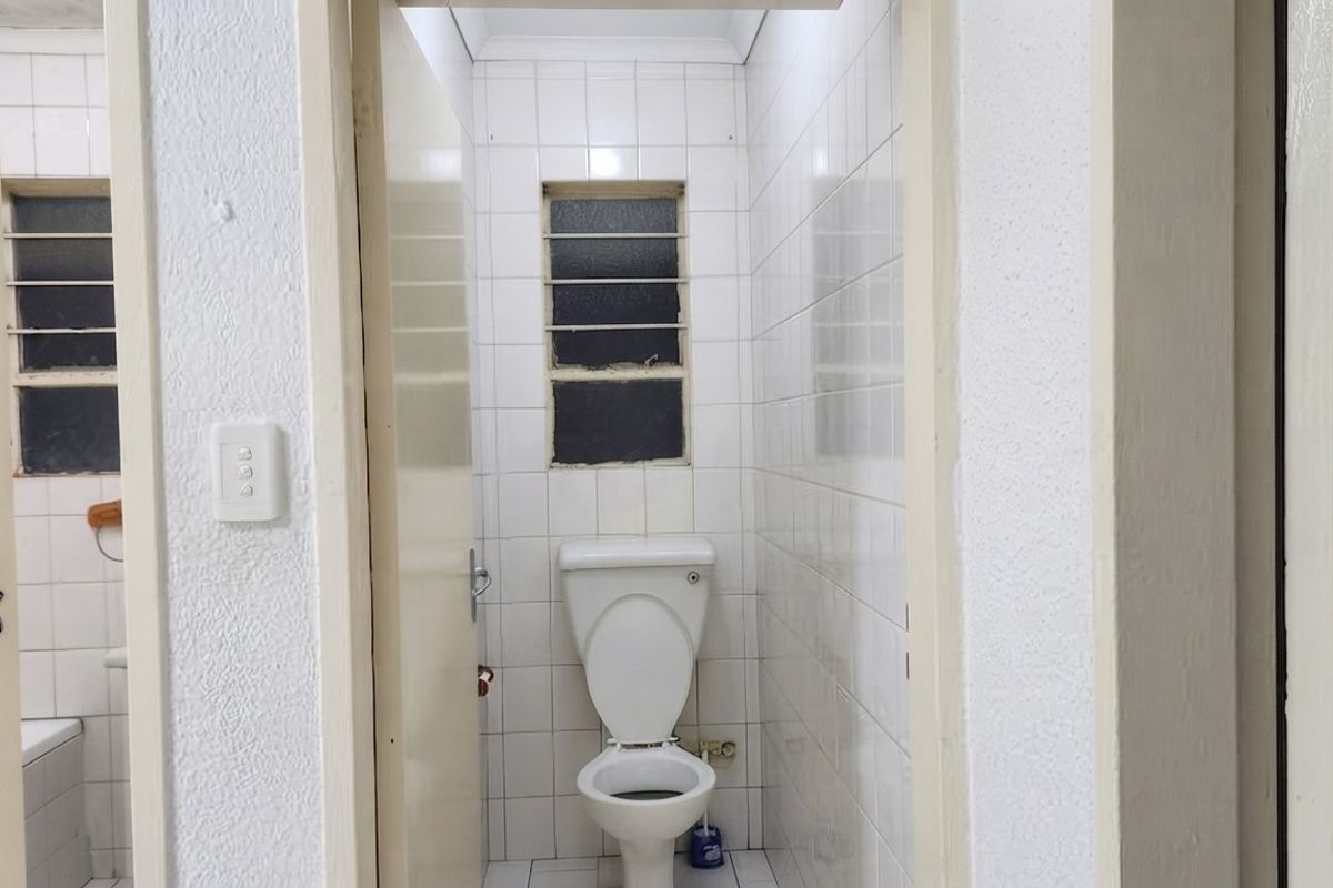 A compact, dated bathroom featuring white wall tiles, a standard toilet, and a window with security bars.