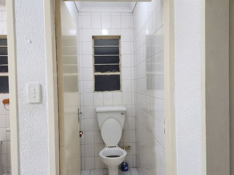 A compact, dated bathroom featuring white wall tiles, a standard toilet, and a window with security bars.
