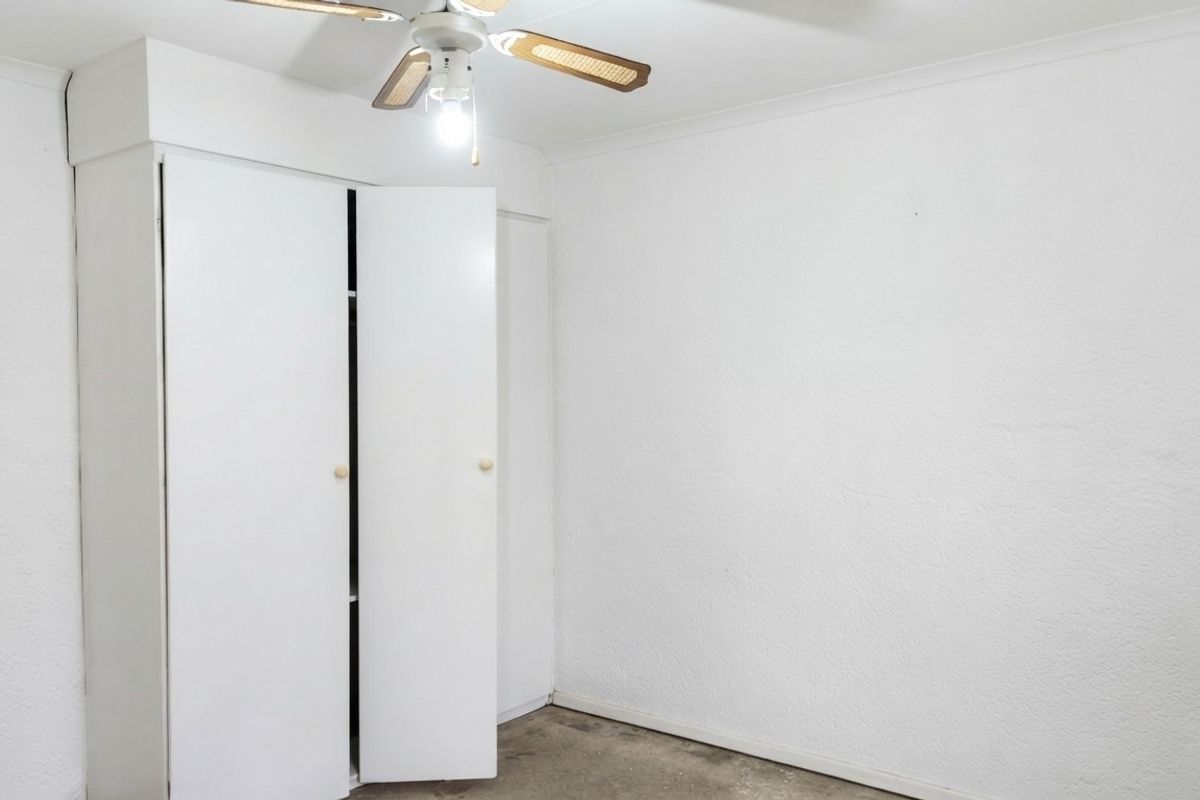 An empty bedroom featuring a built-in wardrobe and a ceiling fan, requiring cosmetic updates to the flooring and walls.