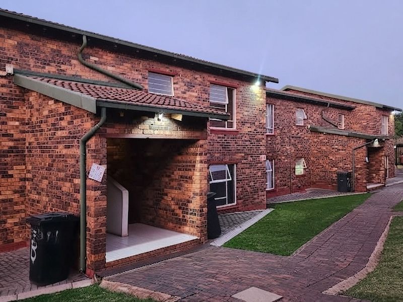 Exterior view of a face-brick townhouse complex featuring paved walkways, tiled roofing, and established garden areas.