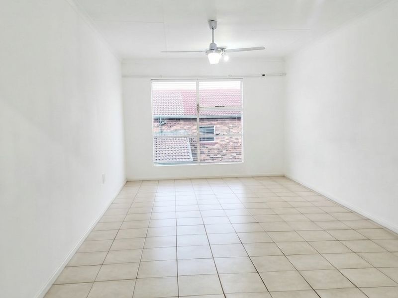 A bright, empty living room featuring neutral tiled flooring, a ceiling fan, and a large window providing natural light.