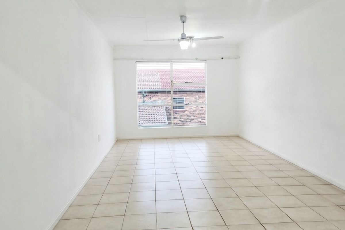 A bright, empty living room featuring neutral tiled flooring, a ceiling fan, and a large window providing natural light.