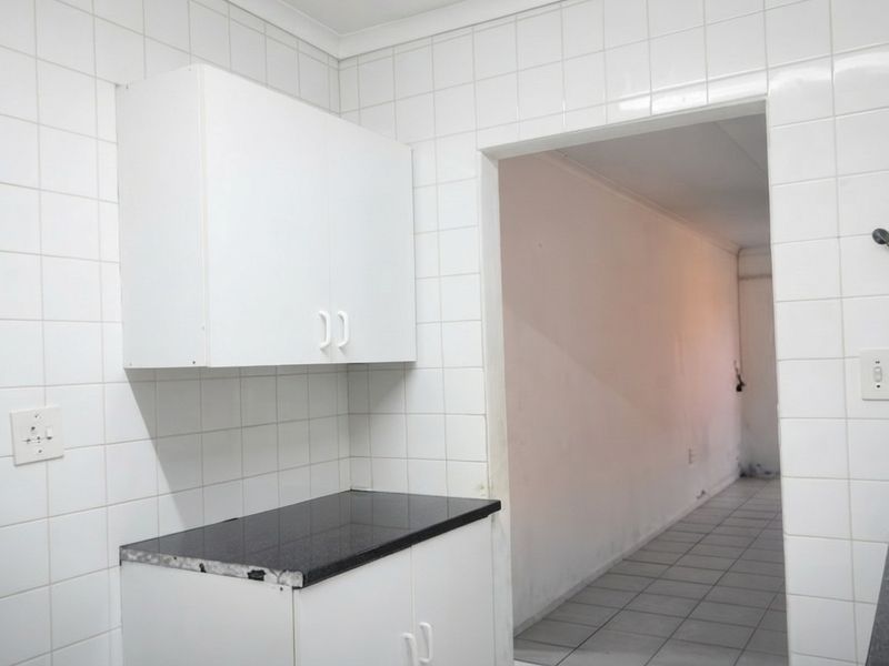 A functional kitchen space featuring white cabinetry, dark granite countertops, and fully tiled walls, leading into an adjacent tiled hallway.