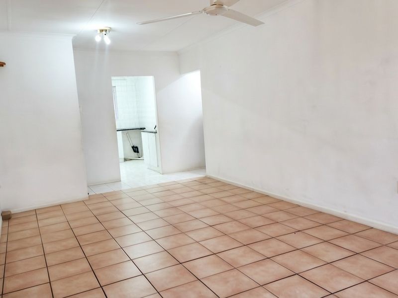 An empty living room featuring ceramic floor tiles, a ceiling fan, and direct access to an adjacent kitchen area.