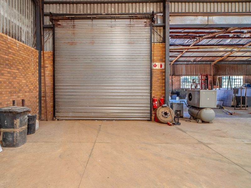 Spacious industrial utility area featuring a large roller shutter door, exposed steel structural beams, and durable concrete flooring within a brick-walled warehouse setting.