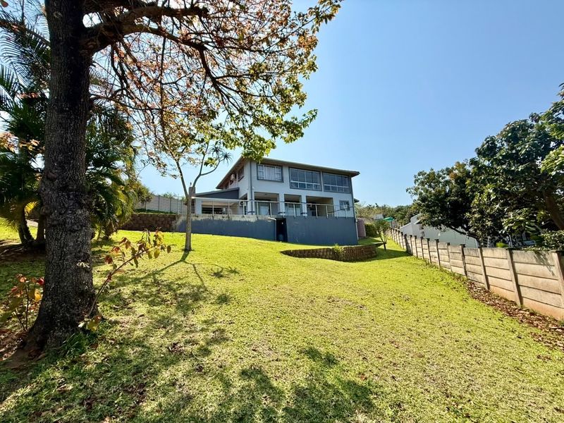 Rear view of a two-story house featuring a spacious lawn, a raised balcony with metal railings, and a concrete retaining wall.