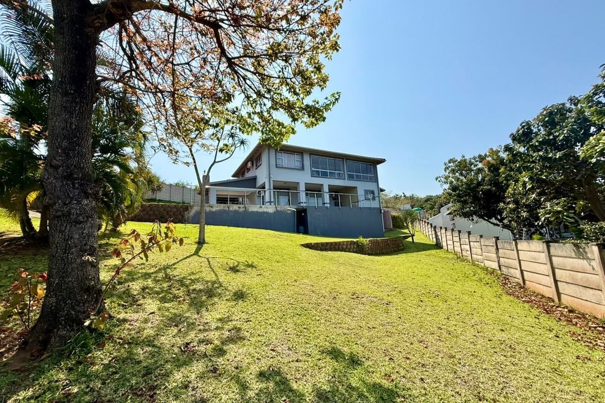 Rear view of a two-story house featuring a spacious lawn, a raised balcony with metal railings, and a concrete retaining wall.