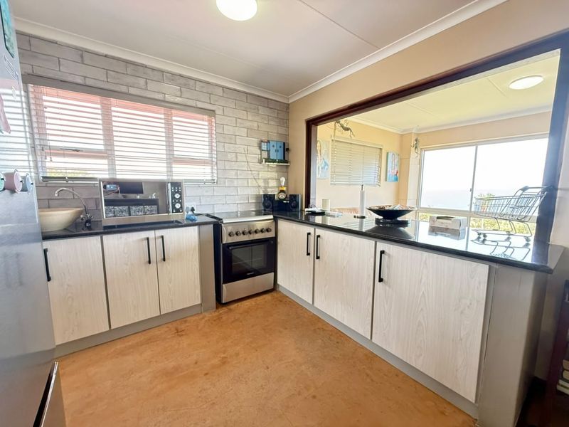 Functional kitchen featuring light wood-grain cabinetry, dark granite countertops, a brick-style feature wall, and a convenient pass-through window overlooking the living area.
