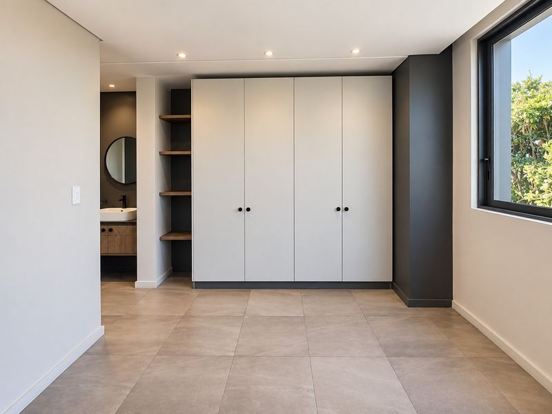 A modern, spacious bedroom featuring neutral tiled flooring, ample built-in cupboard space, recessed shelving, and an integrated en-suite bathroom.