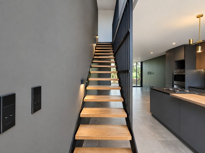 Modern open-plan living area featuring a striking timber staircase with metal balustrade, sleek dark kitchen cabinetry, and high-quality tiled flooring throughout.