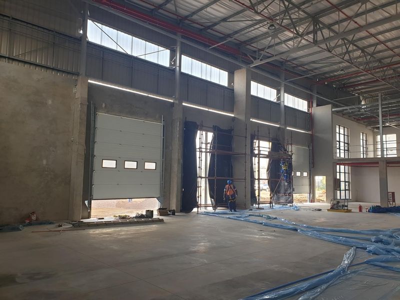Spacious industrial warehouse interior featuring high ceilings with exposed steel trusses, multiple roller shutter doors, and a mezzanine level under construction.