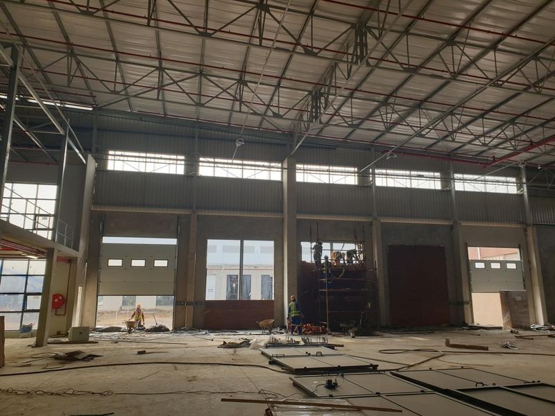 Large industrial warehouse space under construction featuring high ceilings, steel truss roofing, fire sprinkler systems, and multiple roller shutter access doors.