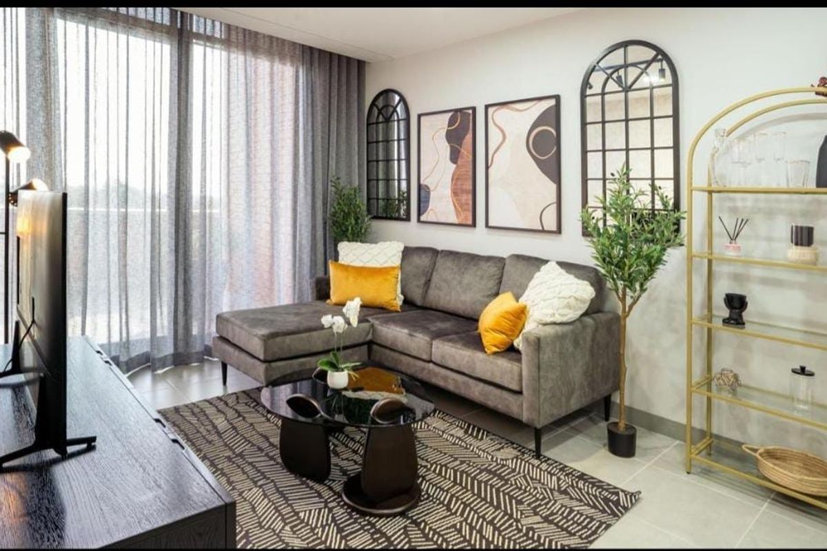 A well-maintained living room featuring large floor-to-ceiling windows with sheer curtains, providing ample natural light and access to the balcony.