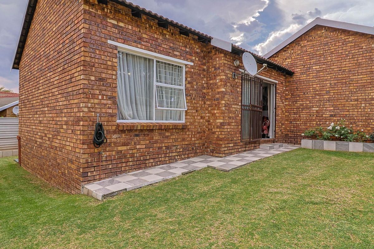 Rear view of a face brick townhouse featuring a small lawn, tiled walkway, and security gate access.