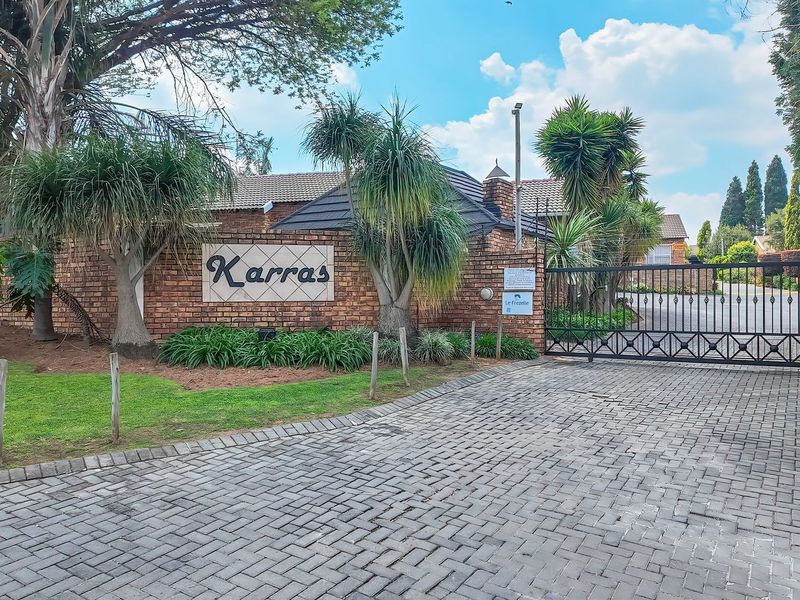 The entrance to Karras townhouse complex featuring a secure metal gate, paved driveway, and established brick boundary wall with landscaped garden.