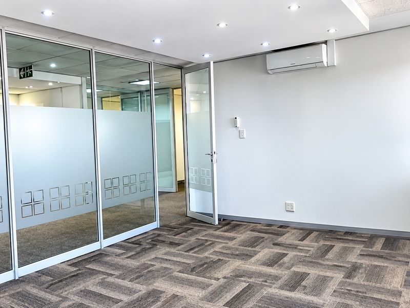 A bright, modern office space featuring glass partition walls, carpet tile flooring, recessed lighting, and a wall-mounted air conditioning unit.