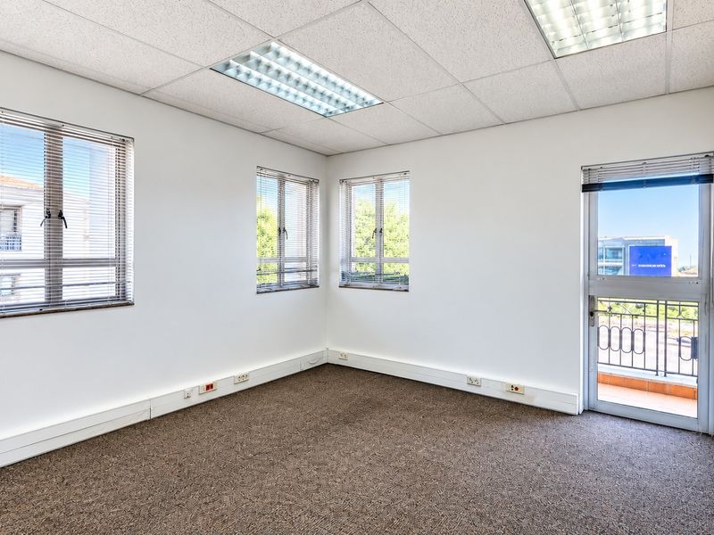 A bright, empty office space featuring carpeted flooring, suspended ceilings with recessed lighting, and direct access to a small balcony.
