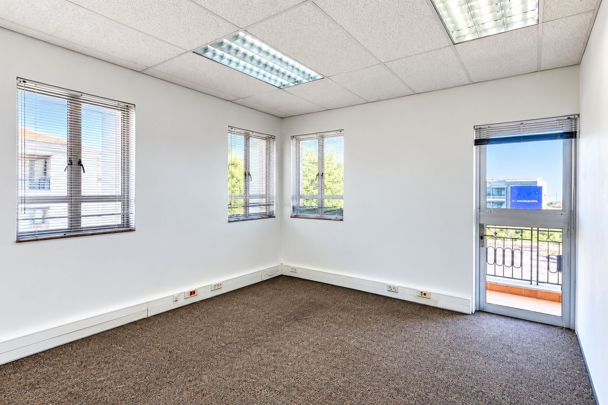 A bright, empty office space featuring carpeted flooring, suspended ceilings with recessed lighting, and direct access to a small balcony.