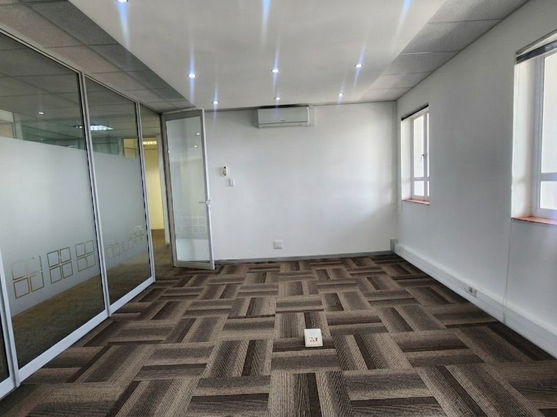 A bright, empty office space featuring modern glass partitions, carpet tile flooring, and a wall-mounted air conditioning unit.