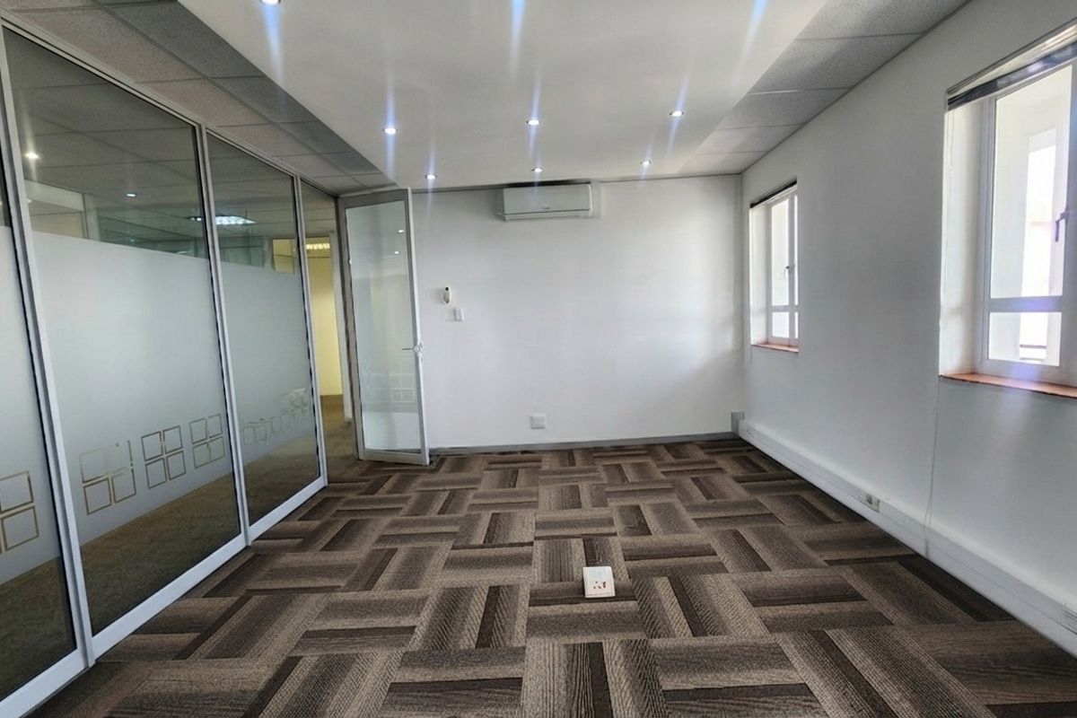 A bright, empty office space featuring modern glass partitions, carpet tile flooring, and a wall-mounted air conditioning unit.