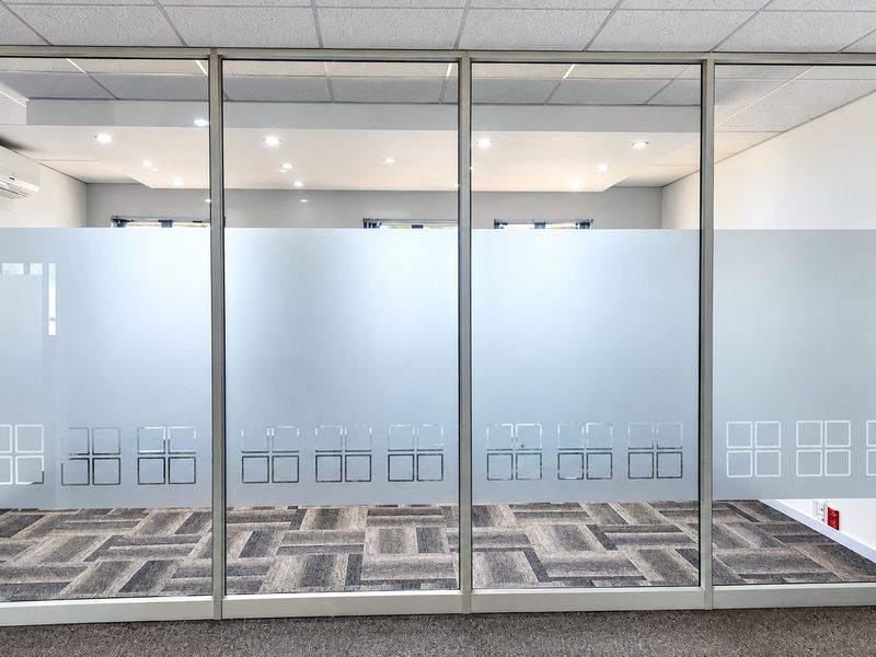 Modern office space featuring a glass partition wall with frosted privacy film, carpeted flooring, and recessed ceiling lighting.