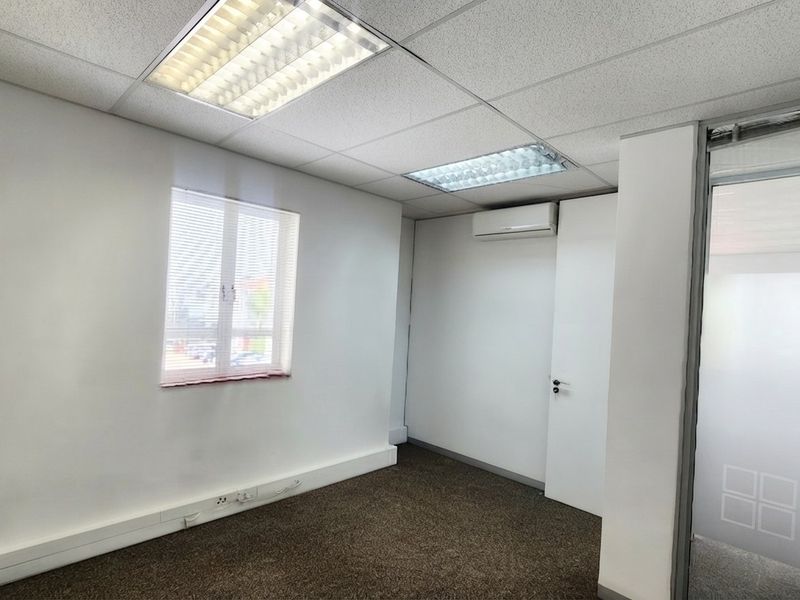 A clean, empty office space featuring carpeted flooring, suspended ceilings with recessed lighting, and a wall-mounted air conditioning unit.