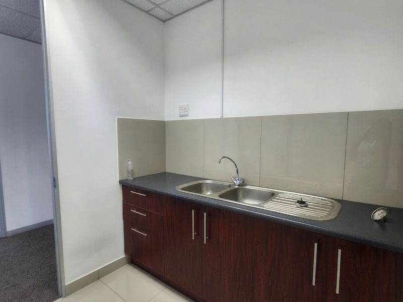 A compact office kitchenette featuring dark wood-finish cabinetry, a stainless steel sink, and neutral tiled splashbacks.