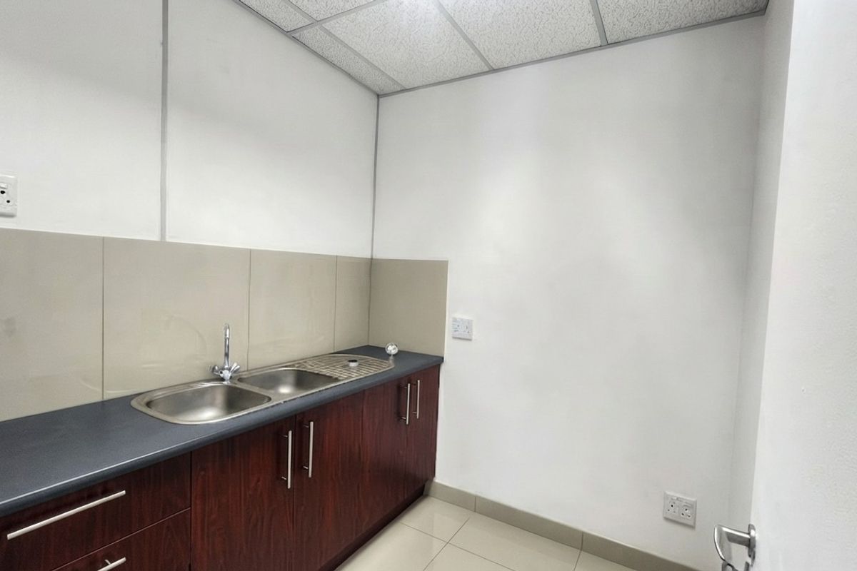 A clean and functional office kitchenette featuring dark wood cabinetry, a stainless steel sink, and neutral tiled finishes.
