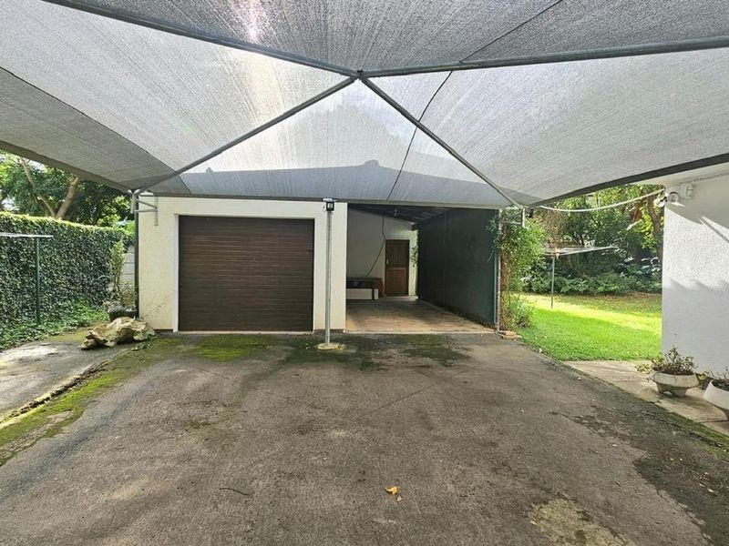 Outdoor parking area featuring a shade cloth carport structure, a roll-up garage door, and a paved driveway with surrounding garden space.