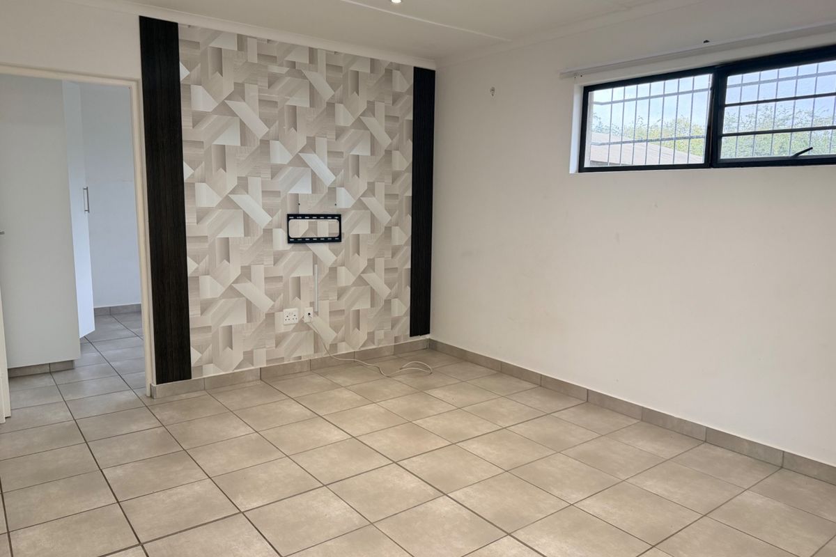 A bright, empty living room featuring neutral tiled flooring, a decorative feature wall, and a window fitted with security bars.